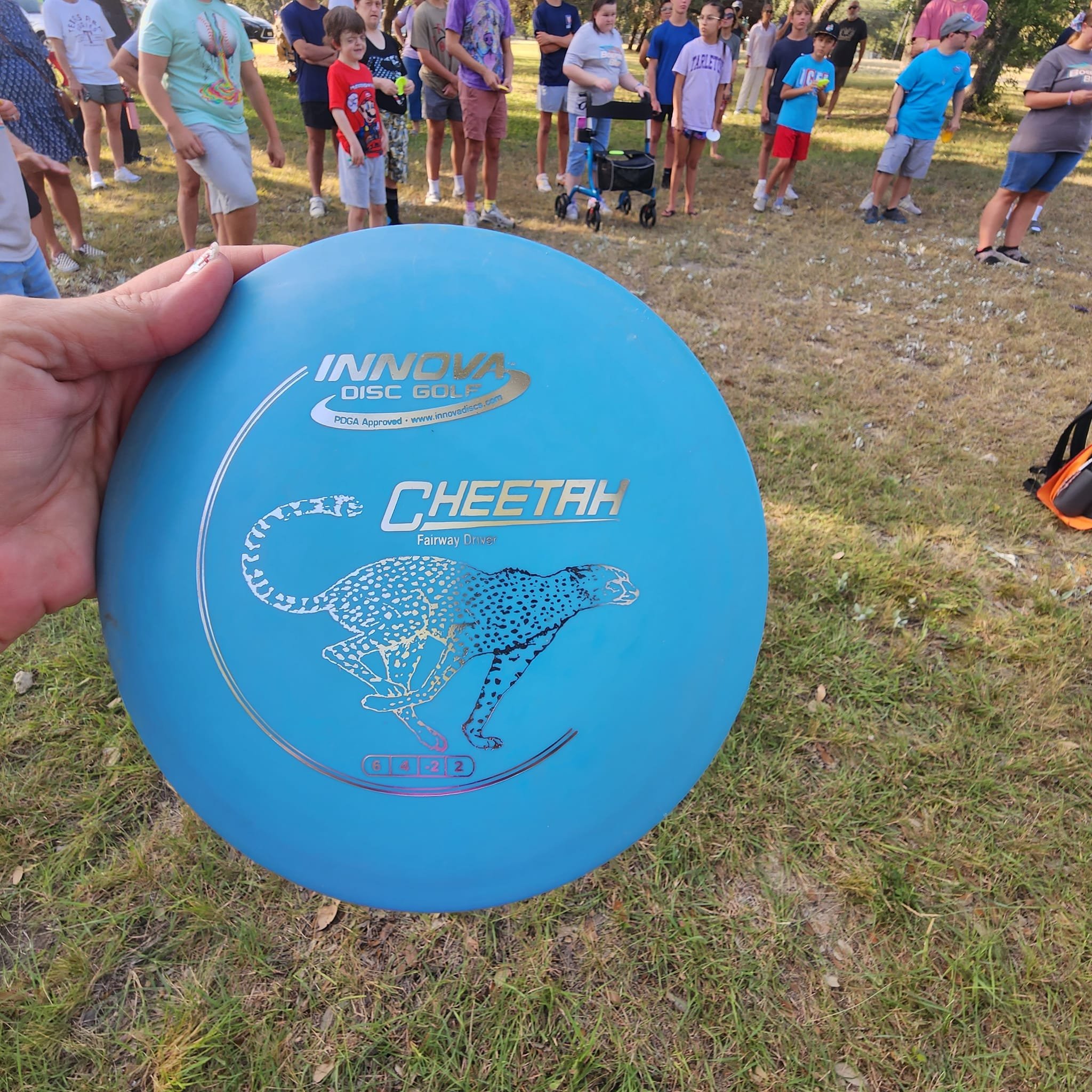 A hand holding a blue disc golf disc with a group of children and adults standing on grass in a park, some with arms crossed and some holding items, with trees in the background.