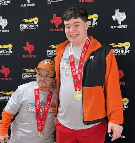 Two young men with medals around their necks pose in front of a black background with logos for Torch Run and Play Unified. One wears a white and orange jacket, and the other wears a white shirt and baseball cap. Both are smiling.