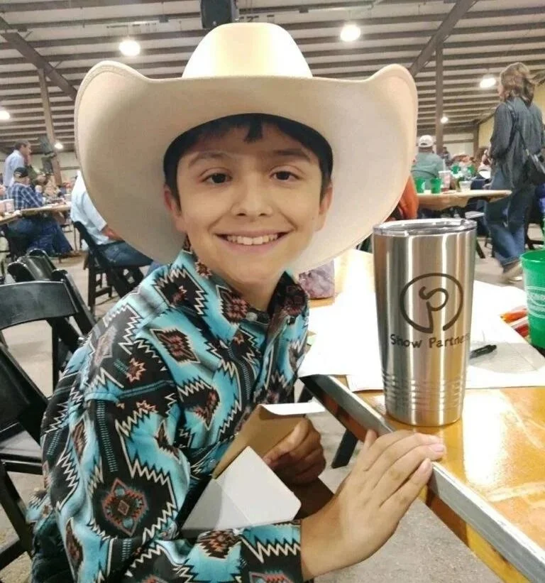 A young boy wearing a cowboy hat and a patterned shirt, smiling at the camera while sitting at a table in a crowded indoor event space.