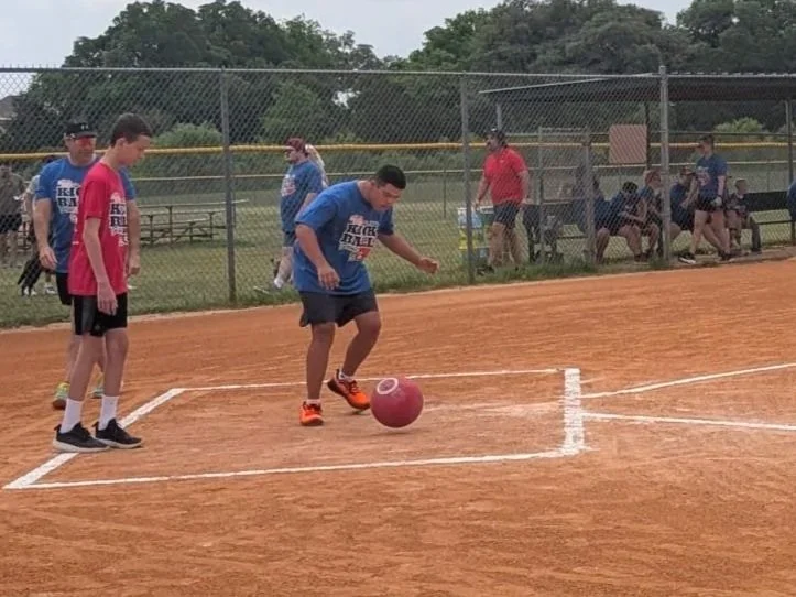 A group of young people on a kickball field, with some standing near the foul line and others sitting on benches behind a chain-link fence, watching during daytime.