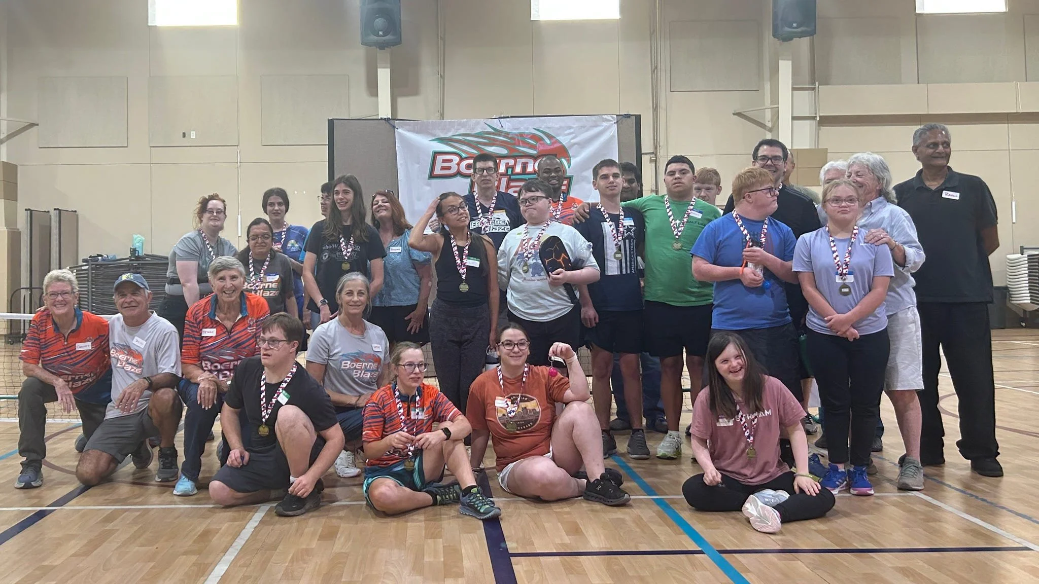 A diverse group of children and adults in a gymnasium, some wearing medals, gathered for a group photo after a sports event, with a banner that reads 'Boerne Blaze' in the background.