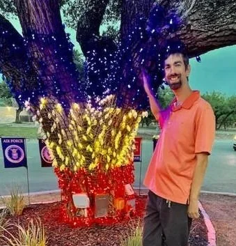A person smiling and standing next to a tree decorated with string lights in red, yellow, and purple colors, resembling a rainbow.