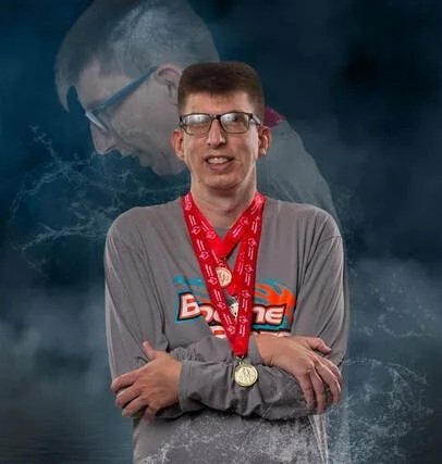 A young man wearing glasses and a gray Langston hoodie, standing with arms crossed, wearing a gold medal around his neck against a dark cloudy background.