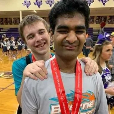 Two smiling young men, one with light skin and the other with darker skin, standing in a gymnasium, with the man in the front wearing a medal around his neck and the person behind him resting their hands on his shoulders.