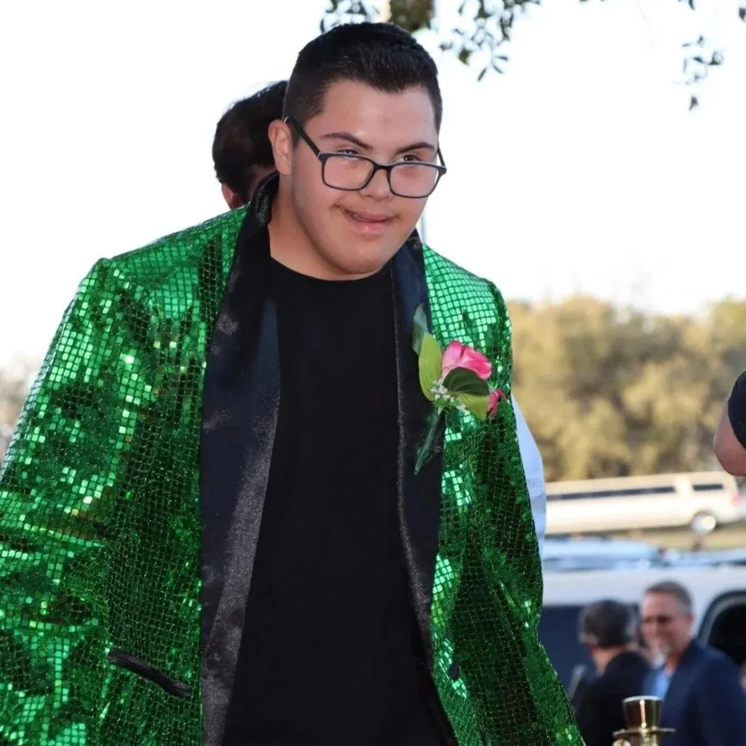 A young man wearing a sparkly green jacket and glasses, with a pink boutonniere on his left lapel, smiling at an outdoor event.
