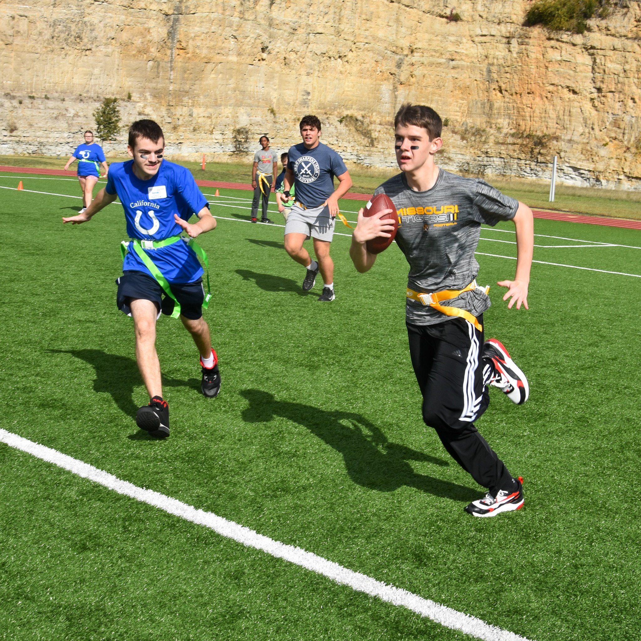 Young boys and a girl running during a football game on a grassy field, with a rocky cliff in the background.