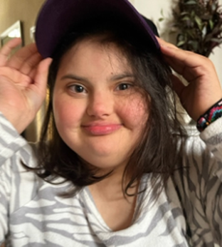 A young girl with dark hair and a light complexion wearing a gray and white striped shirt, holding a purple hat on her head, smiling at the camera.