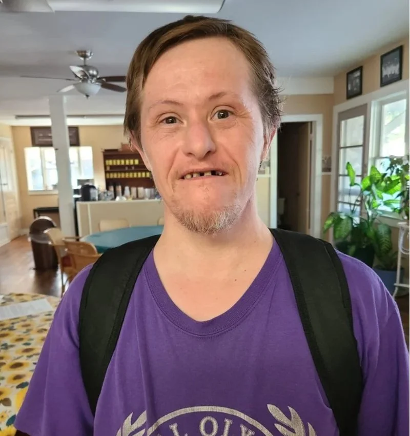 A man with brown hair, missing teeth, and a goatee, wearing a purple T-shirt and carrying a black backpack, standing in a sunlit living room with plants and a dining area in the background.