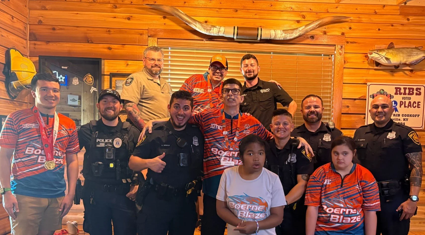 Group photo of young cyclists, police officers, and individuals in a wood-paneled room, some wearing medals and jerseys with 'Boerne Blaze' logos, with a mounted fish and a ribcooked sign in the background.