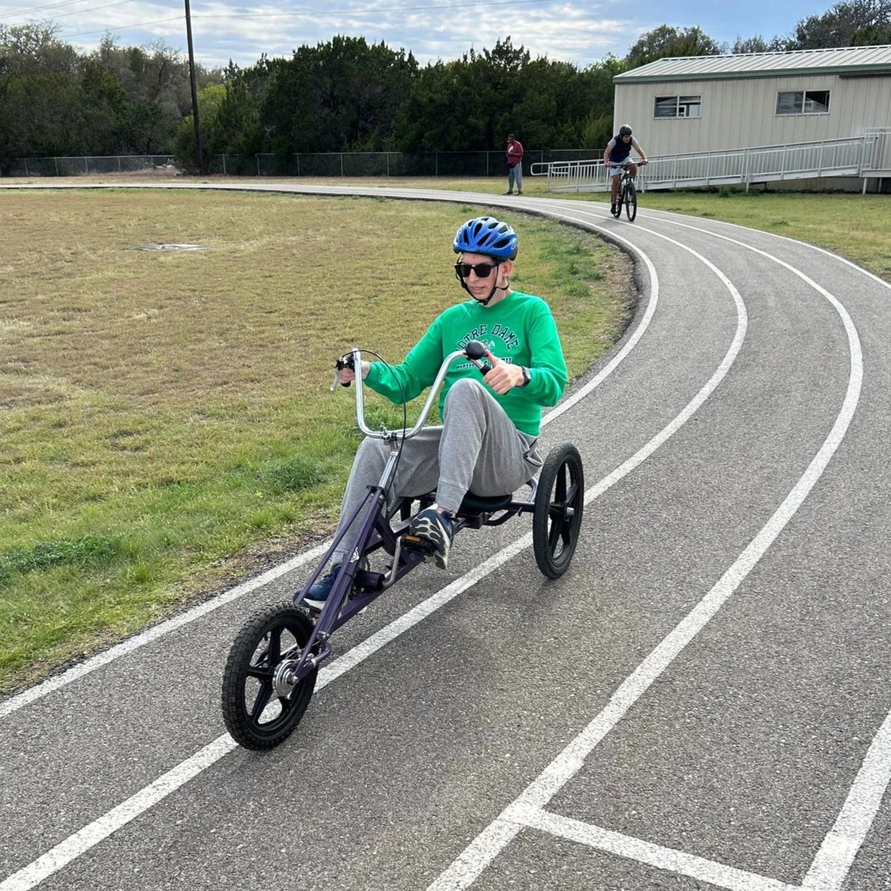 Person riding a recumbent tricycle on a curved outdoor track, wearing a blue helmet, sunglasses, and a green sweatshirt, with two other cyclists and a person standing in the background.