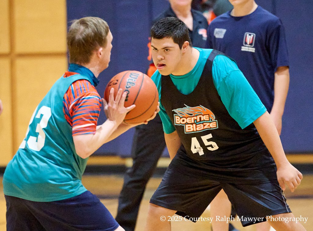 Two young male basketball players face off during a game, with one holding the basketball and the other in a defensive stance. The player with the ball wears a teal and orange jersey numbered 13, while the defender wears a black jersey with teal sleeves numbered 45 and the logo 'Boerne Blaze'.