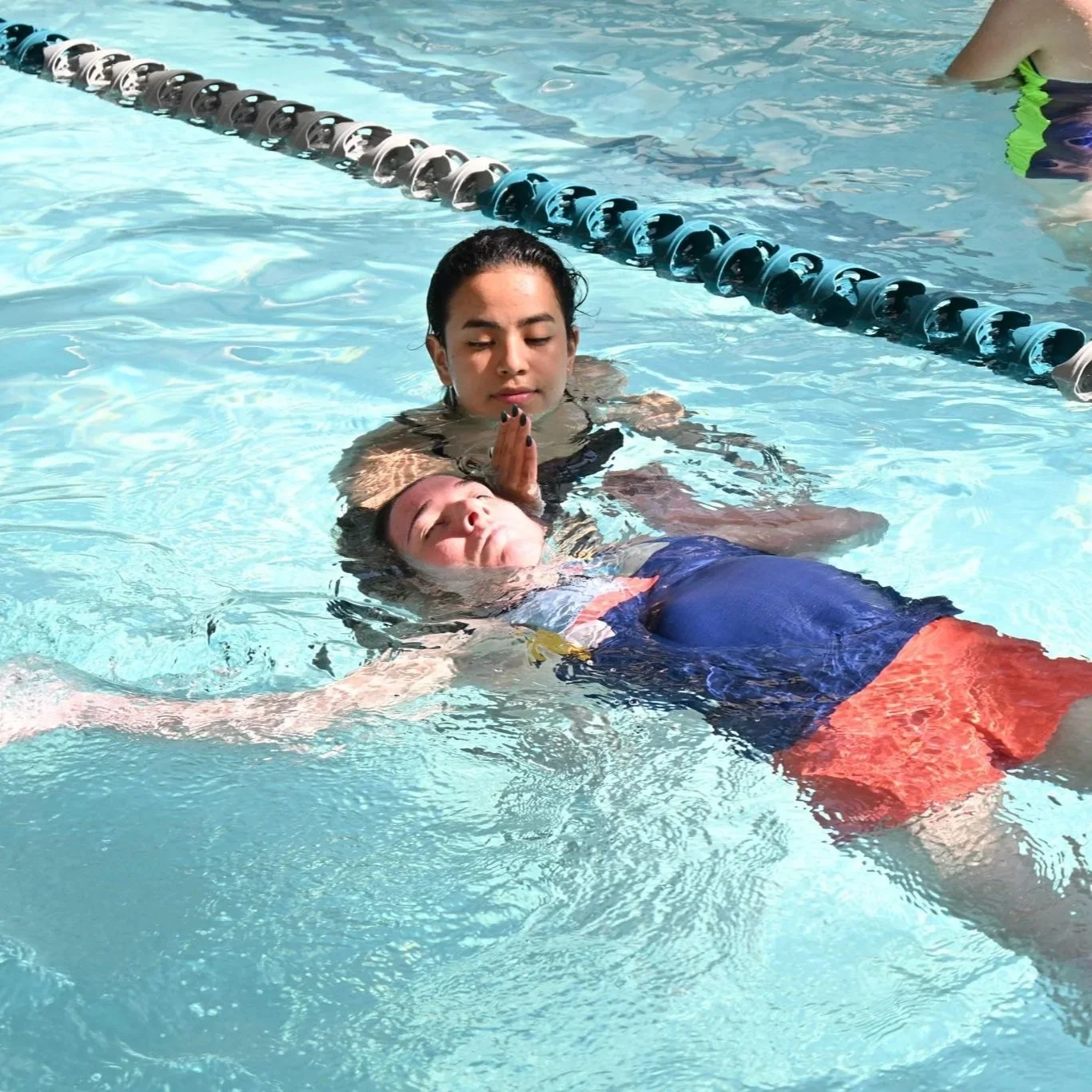 A woman appears to be performing a rescue in a swimming pool, holding a young child underwater with her hand placed gently on the child's head. The child is wearing a cap and colorful swimsuit, while the woman is in a black swimsuit. There is a lane marker in the pool, and part of another person can be seen in the background.