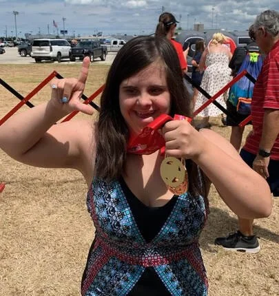 A young girl smiling and holding a red phone, making a hand sign with her fingers, at an outdoor event with people and cars in the background.