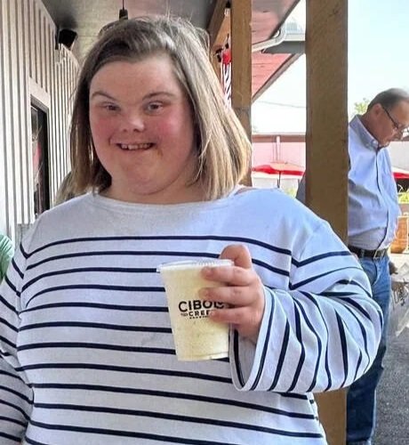 A smiling woman holding a cup of shaved ice outdoors with a man in the background.
