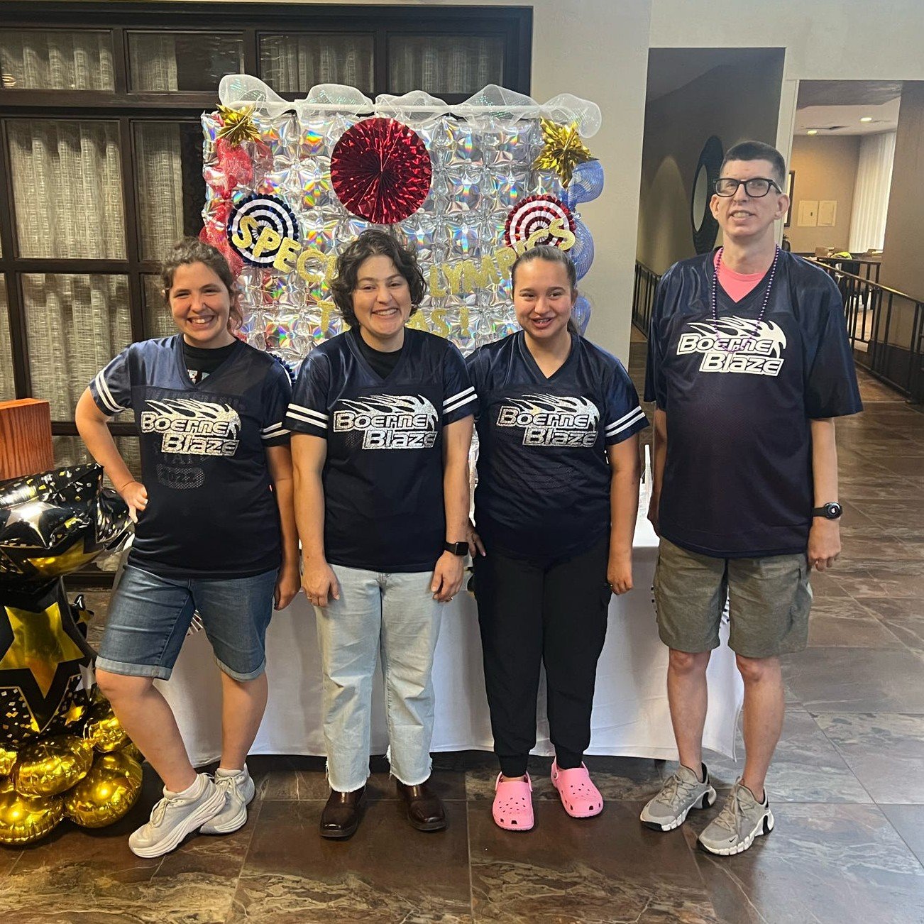 Four people wearing matching black 'Boeing Blaze' jerseys posing indoors in front of a party backdrop decorated with balloons and foil decorations, celebrating a special occasion.