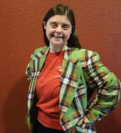 Young woman smiling, with dark brown hair, wearing a red shirt and a green plaid blazer, standing against a reddish-brown textured wall.