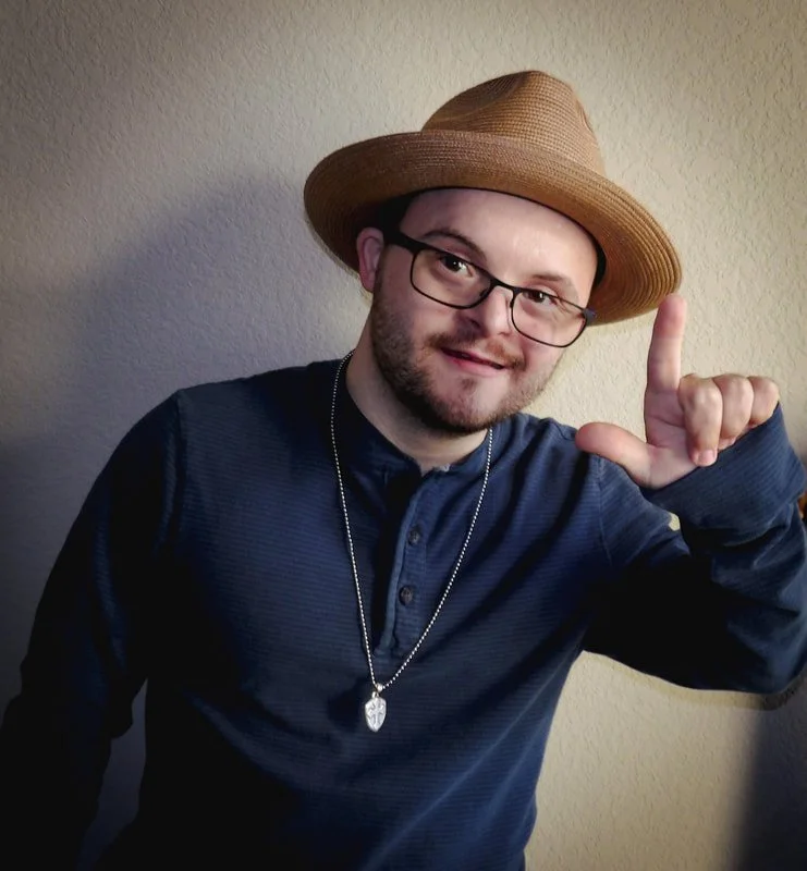A young man with glasses, a beard, and a mustache wearing a tan straw hat, a dark blue button-up shirt, and a silver chain necklace with a leaf pendant, making an 'L' shape with his right hand.