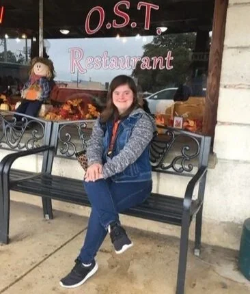 A young woman sitting on a black bench outside of a restaurant with a doll in the window behind her, which has a sign reading 'O.S.T Restaurant'.