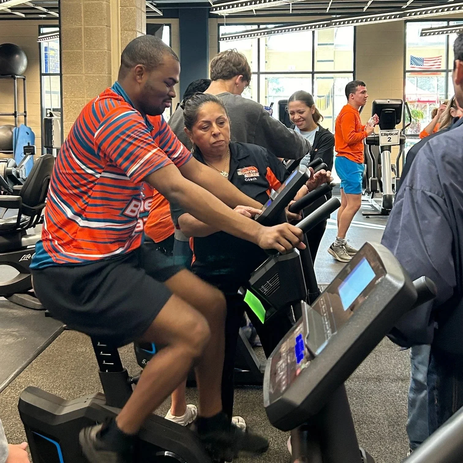 People exercising on indoor stationary bikes at a gym, with a coach monitoring.