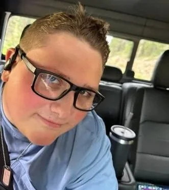 A young man wearing glasses and a blue shirt sitting in the backseat of a car, smiling at the camera.
