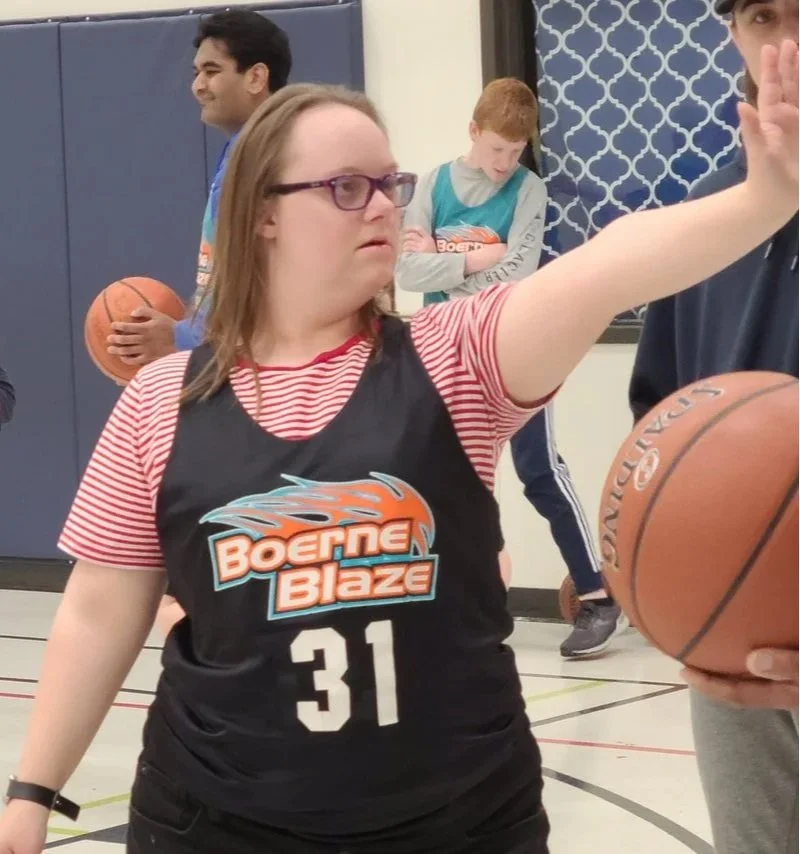 A woman in a black basketball jersey with the number 31 and 'Boerne Blaze' logo is playing basketball indoors. Other children are in the background, one holding a basketball and another looking down.