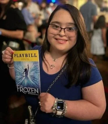 A smiling girl with glasses and long dark hair holding a Disney Frozen playbill at an indoor event.