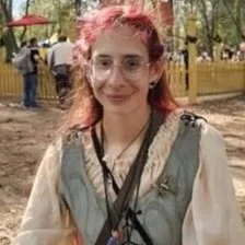 A young woman with glasses and pink hair, smiling outdoors in a park setting.
