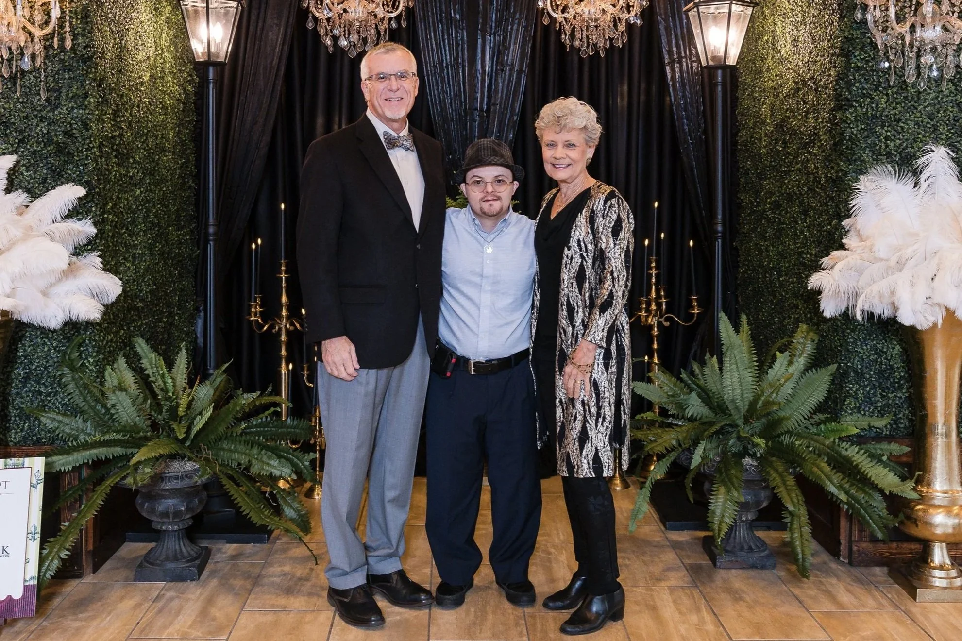 Three people standing in front of a decorated backdrop with black curtains, chandeliers, and greenery. They are well-dressed, smiling, with the person in the middle wearing glasses and a hat.