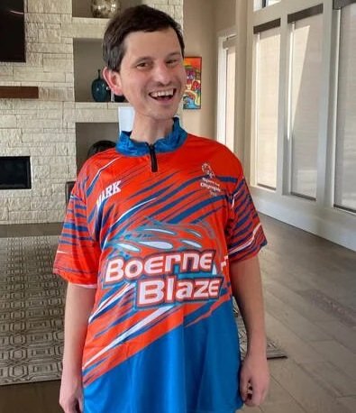 A young man with short dark hair smiling, wearing a red and blue sports jersey with the text 'Boeing Blaze,' standing indoors in front of large windows.