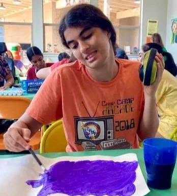 A young girl painting with purple paint on a white surface, holding a paintbrush in her right hand and a green and black object in her left hand, seated at a table in a classroom with other children in the background.