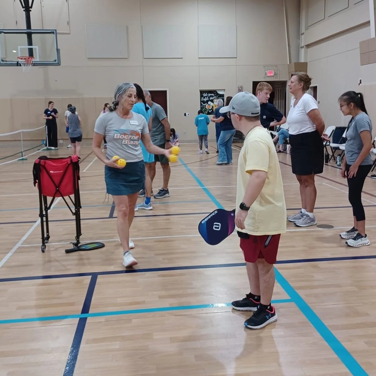 A woman and a young boy playing pickleball in an indoor gym. The woman is holding yellow pickleballs, and the boy is holding a pickleball paddle. Several other people are in the background participating in or observing the activity.