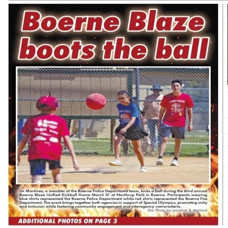 Children playing kickball during a community event, with a chain-link fence in the background and a red kickball in mid-air.