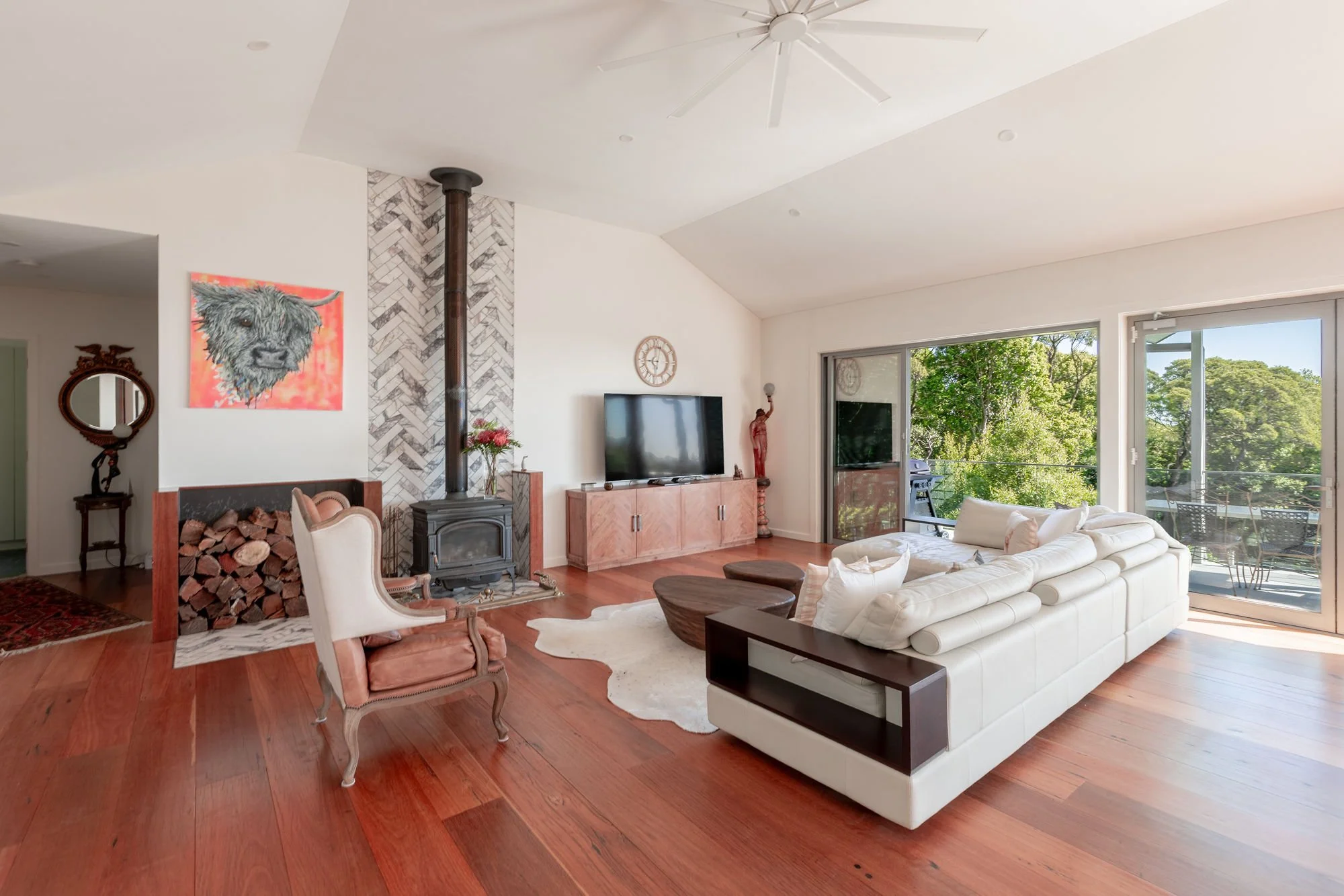 Living room with a white sofa, wooden chairs, a wood-burning stove, a large window showing greenery outside, a ceiling fan, and various decorative items including a painting of a dog and a wall clock.
