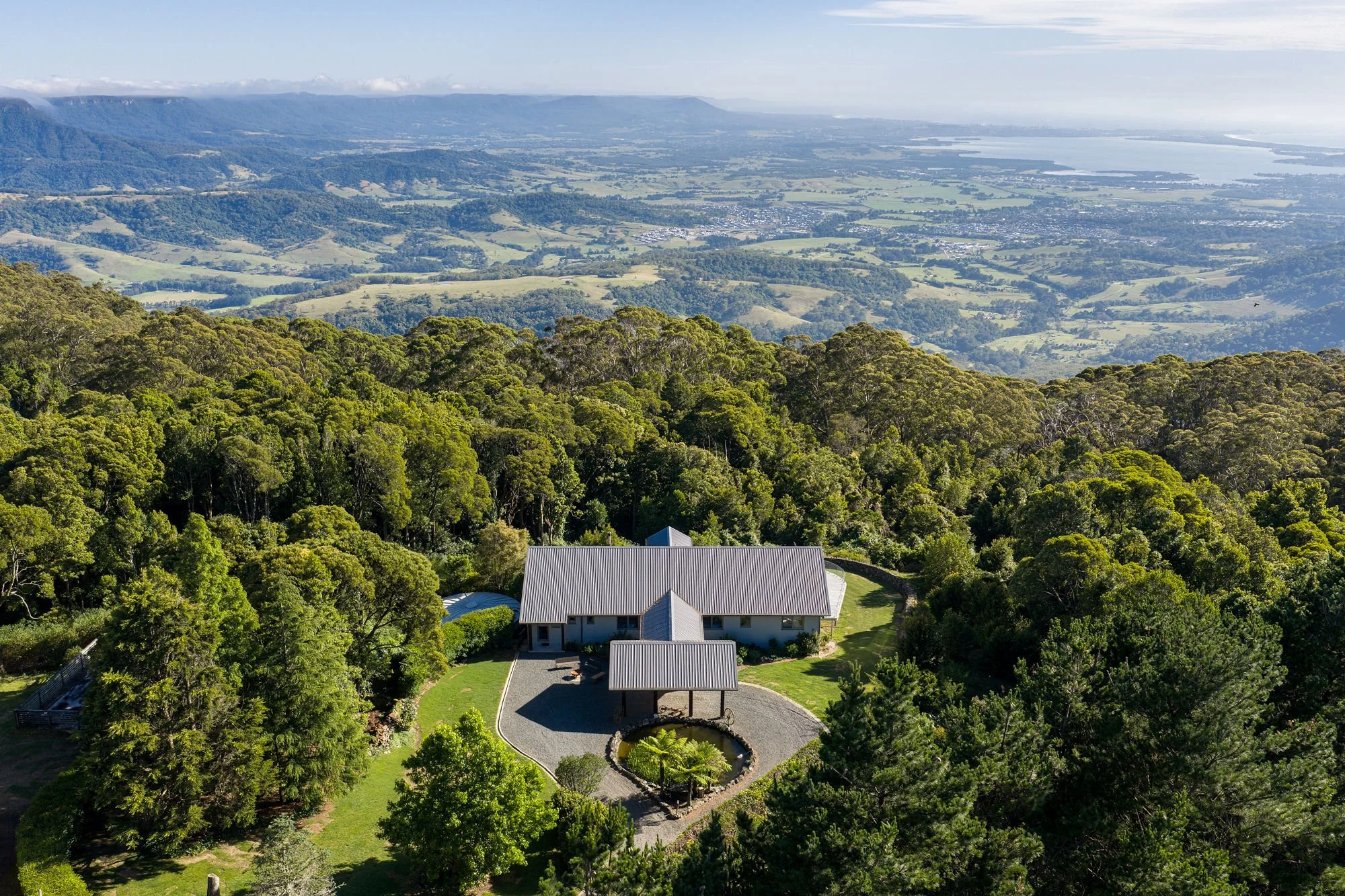 A house with a metal roof surrounded by lush trees and a well-maintained garden, overlooking rolling hills and distant bodies of water.