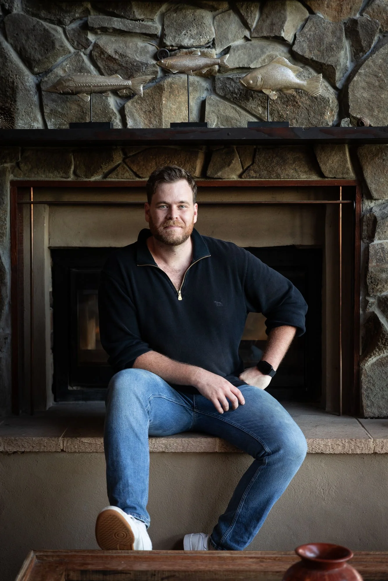 A man sitting in front of a stone fireplace with decorative fish figurines mounted above the mantel. The man has short dark hair, a beard, and is wearing a black zip-up sweatshirt and blue jeans. He has his left hand on his hip and his right arm resting on his knee. The fireplace is surrounded by large stones, and the photo captures him from the waist up, sitting on the hearth.
