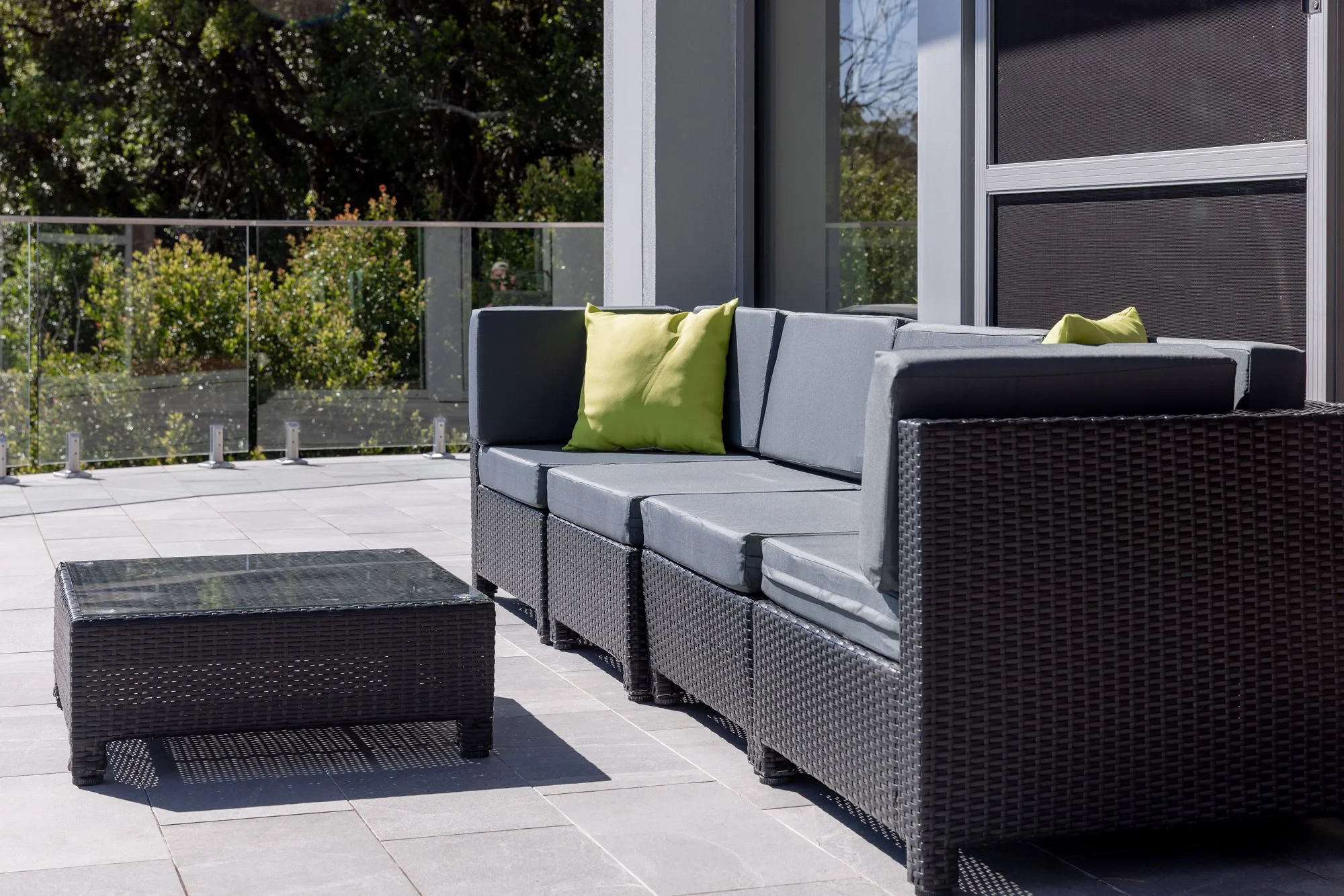 Outdoor patio with gray wicker furniture including a sectional sofa with green pillows, a coffee table, and a glass-topped side table. The patio has gray tiled flooring, a glass railing, and lush green foliage in the background.