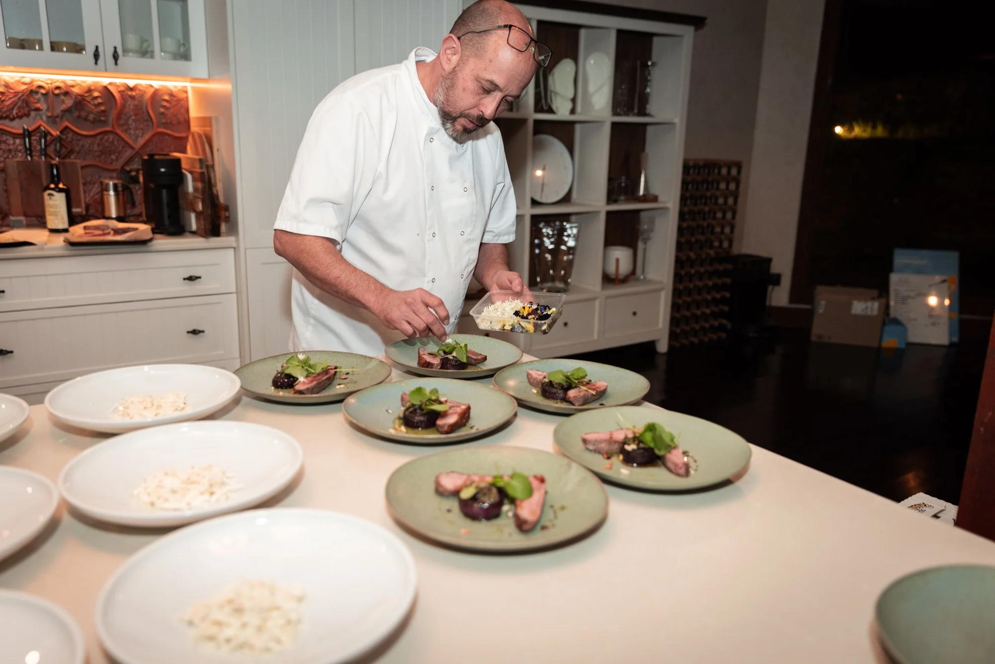 A chef in a white chef coat prepares multiple plates of a gourmet dish in a professional kitchen with shelves and kitchen tools in the background.