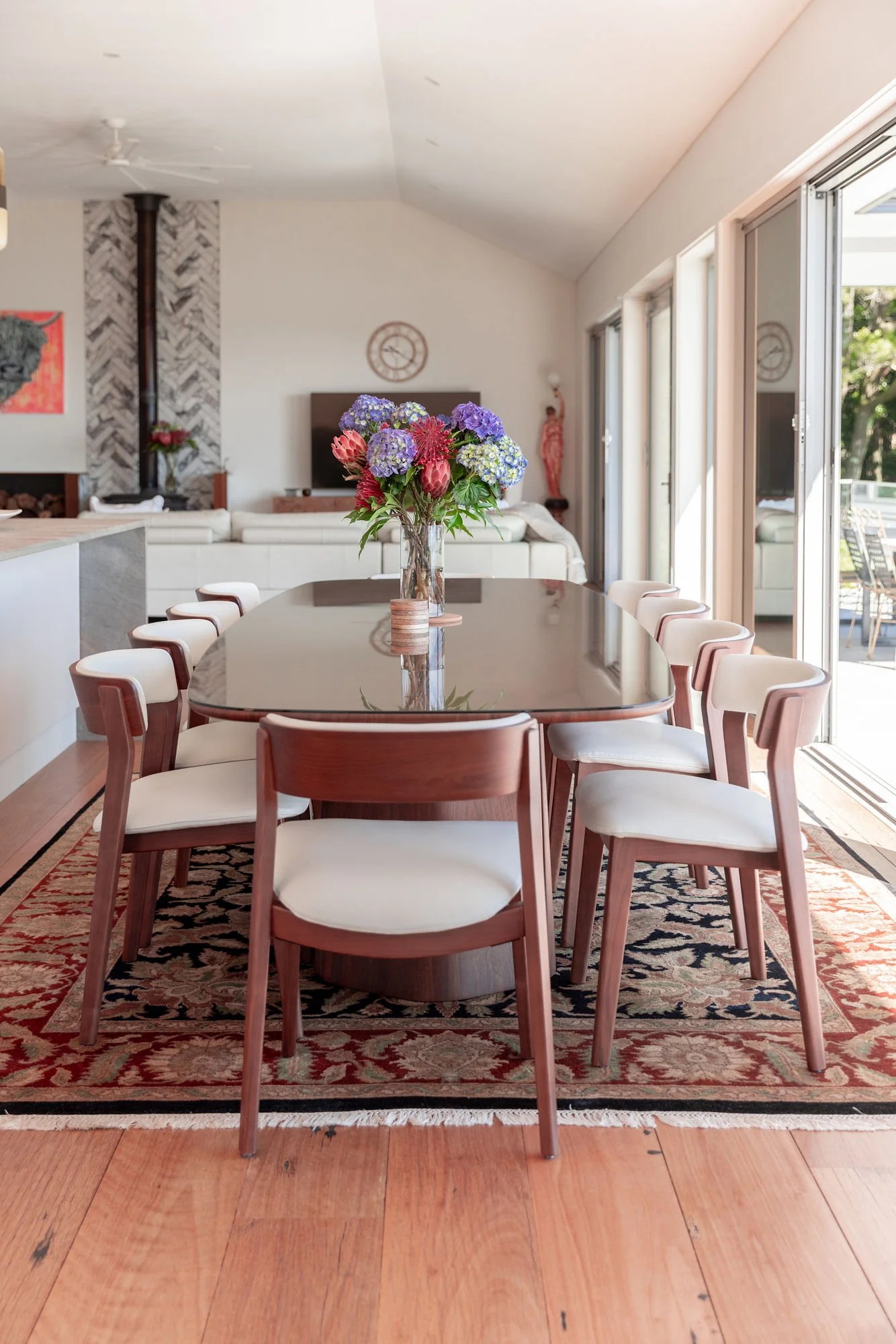 Dining room with a long wooden table and white cushioned chairs, a vase with purple and pink flowers, large sliding glass doors leading to an outdoor patio, a patterned rug beneath the table, and artwork on the walls.