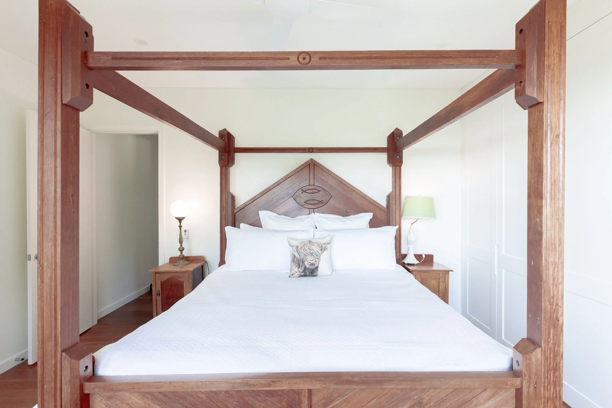 A bedroom with a four-poster bed made of wood and white linens, with a decorative pillow featuring a dog, on a wooden floor. Two nightstands with lamps are on either side of the bed.