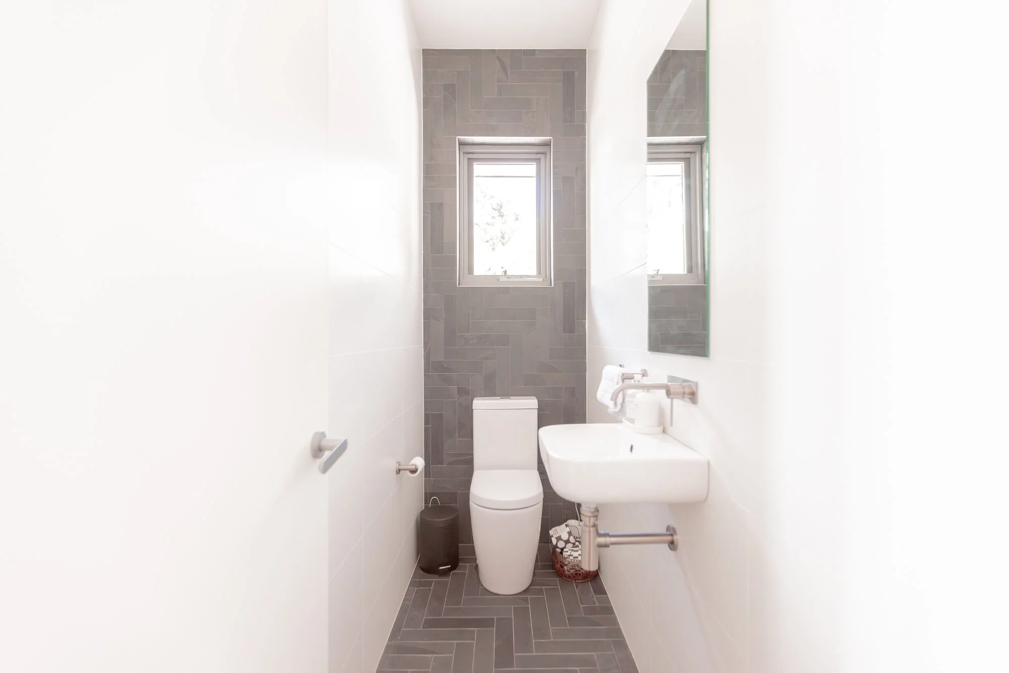 Small modern bathroom with gray tiled wall behind toilet, white sink, and two high small windows.
