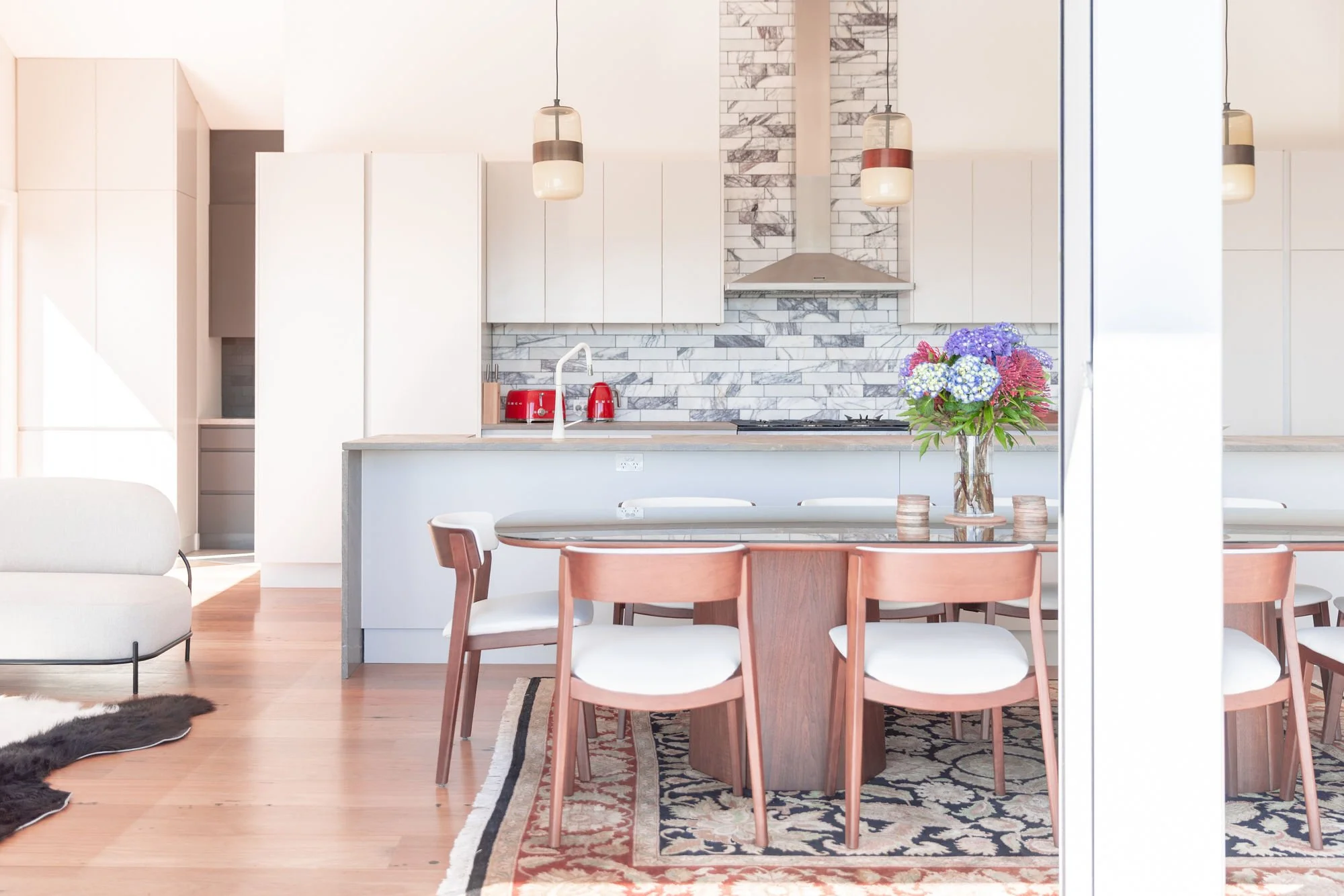 Modern kitchen with white cabinetry, marble backsplash, and stainless steel hood, adjacent to a dining area with wooden chairs and a glass vase of colorful hydrangeas on a rug.