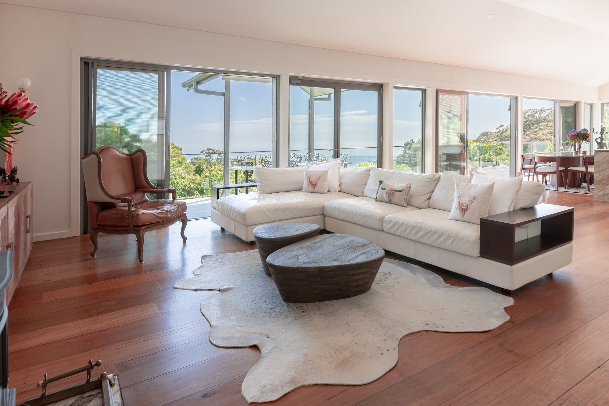Living room with white sectional sofa, wooden coffee tables, armchair, and floor-to-ceiling windows showing trees and blue sky outside.