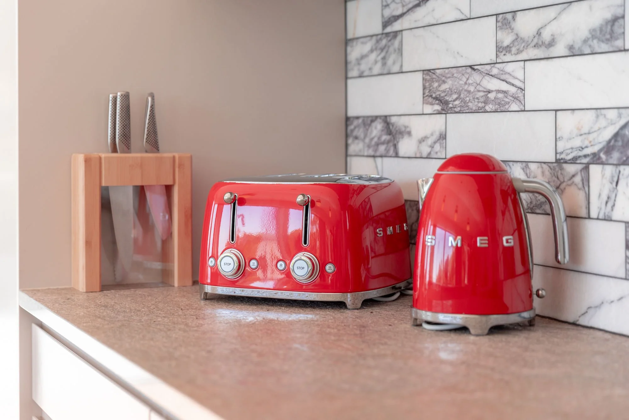 Red SMEG toaster and kettle on a kitchen countertop with a marble tile backsplash and a wooden knife holder with knives.