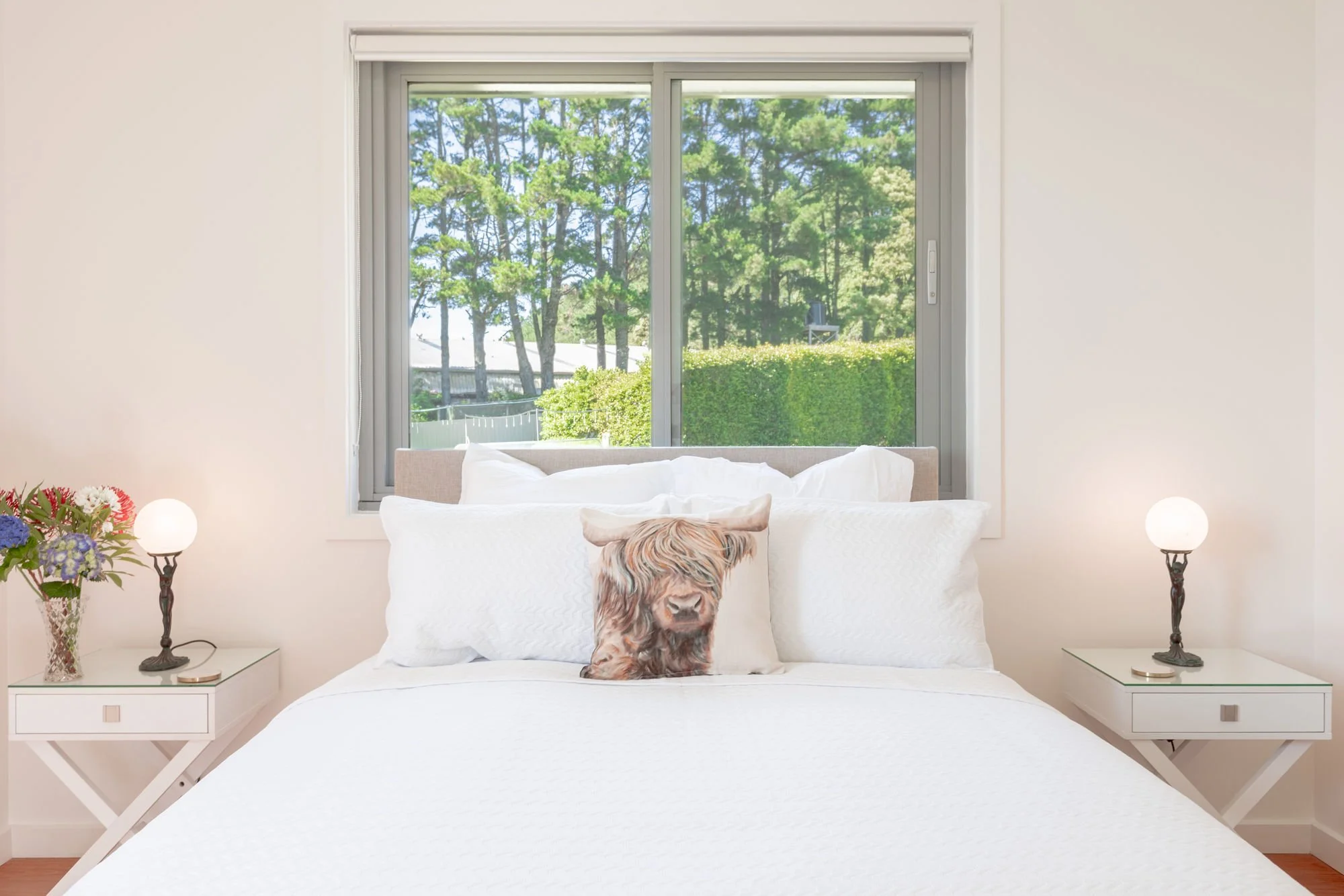 A bedroom with a large window showing green trees outside. The bed has white bedding and a pillow with a dog face. There are two nightstands with lamps and a vase with flowers.