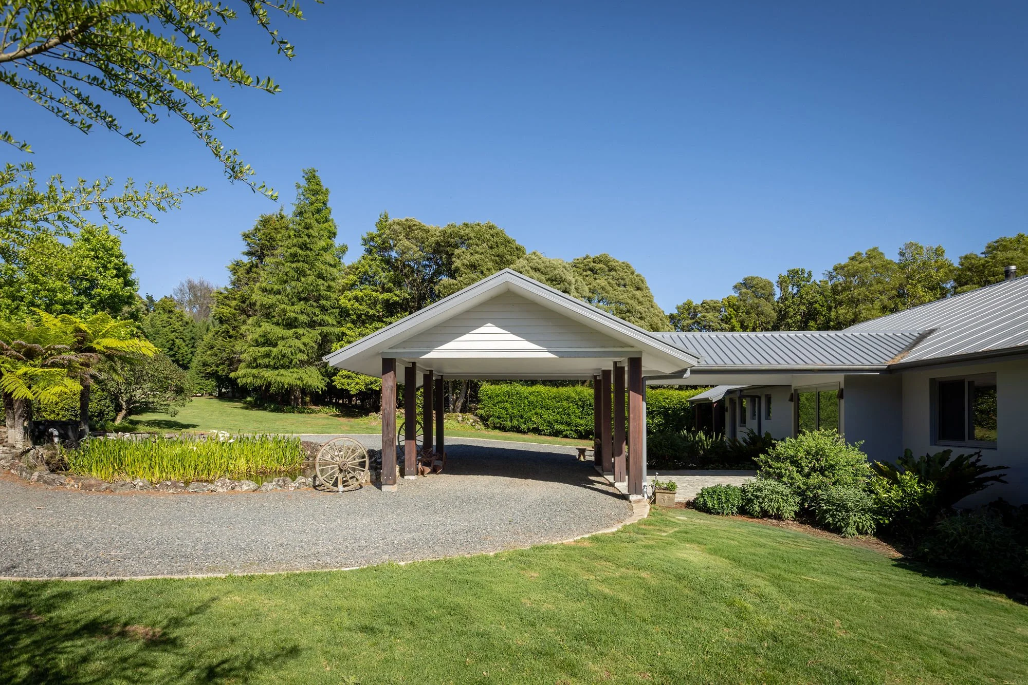 A house with a silver metal roof and white siding, surrounded by lush green trees and grass under a clear blue sky, with a small garden and a gravel pathway in the foreground.