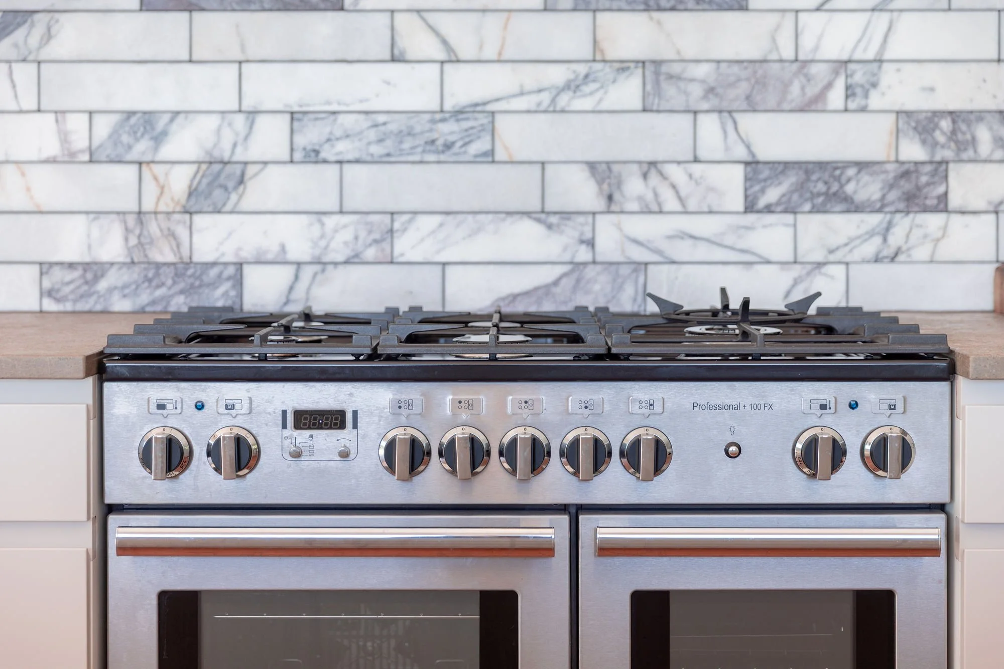 A stainless steel professional gas stove with six burners, control knobs, and a digital clock, installed in a kitchen with a marble tile backsplash and beige cabinetry.