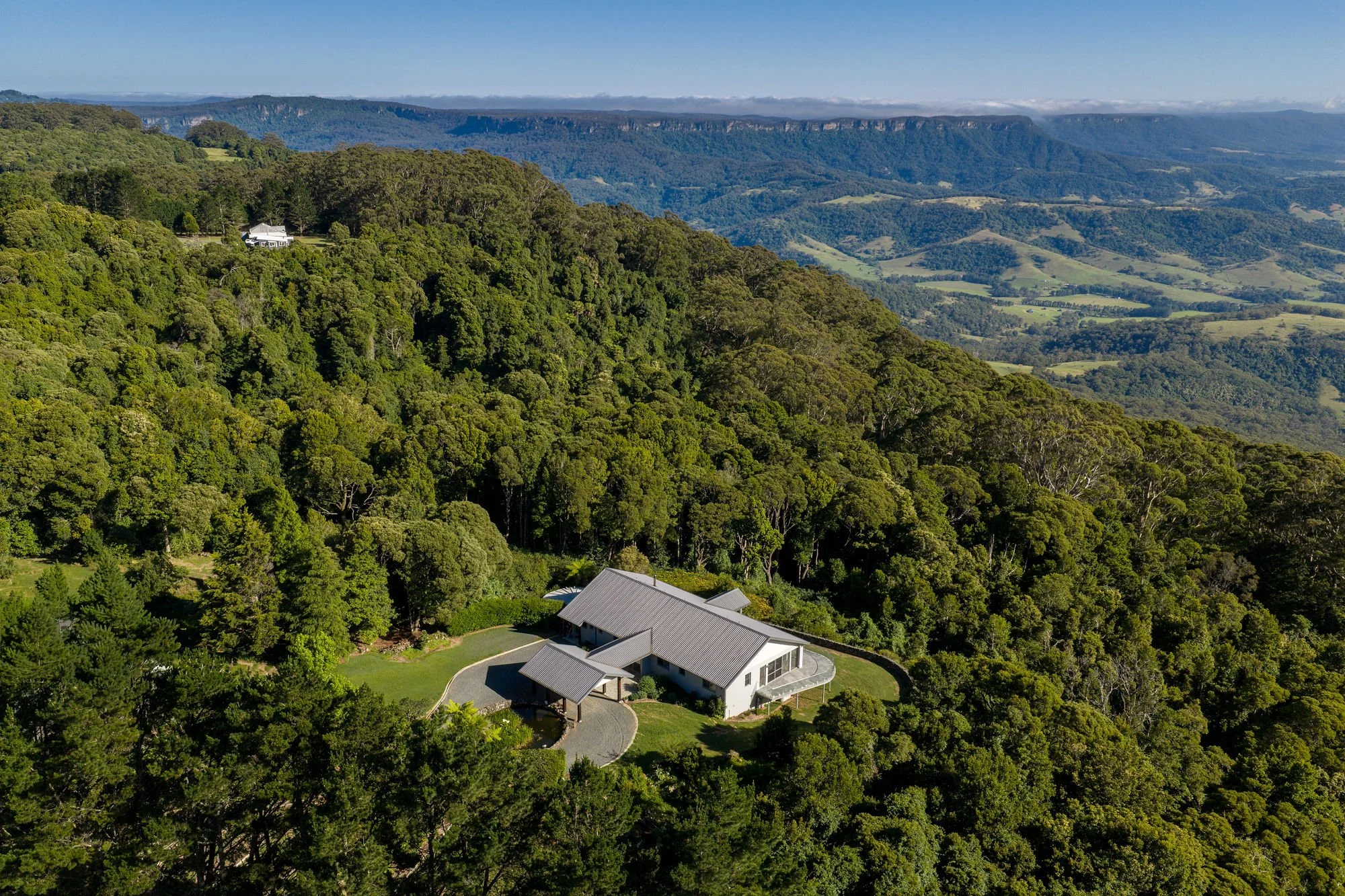 Aerial view of a house surrounded by dense green forest on a hillside, with rolling hills and mountains in the background under a clear blue sky.
