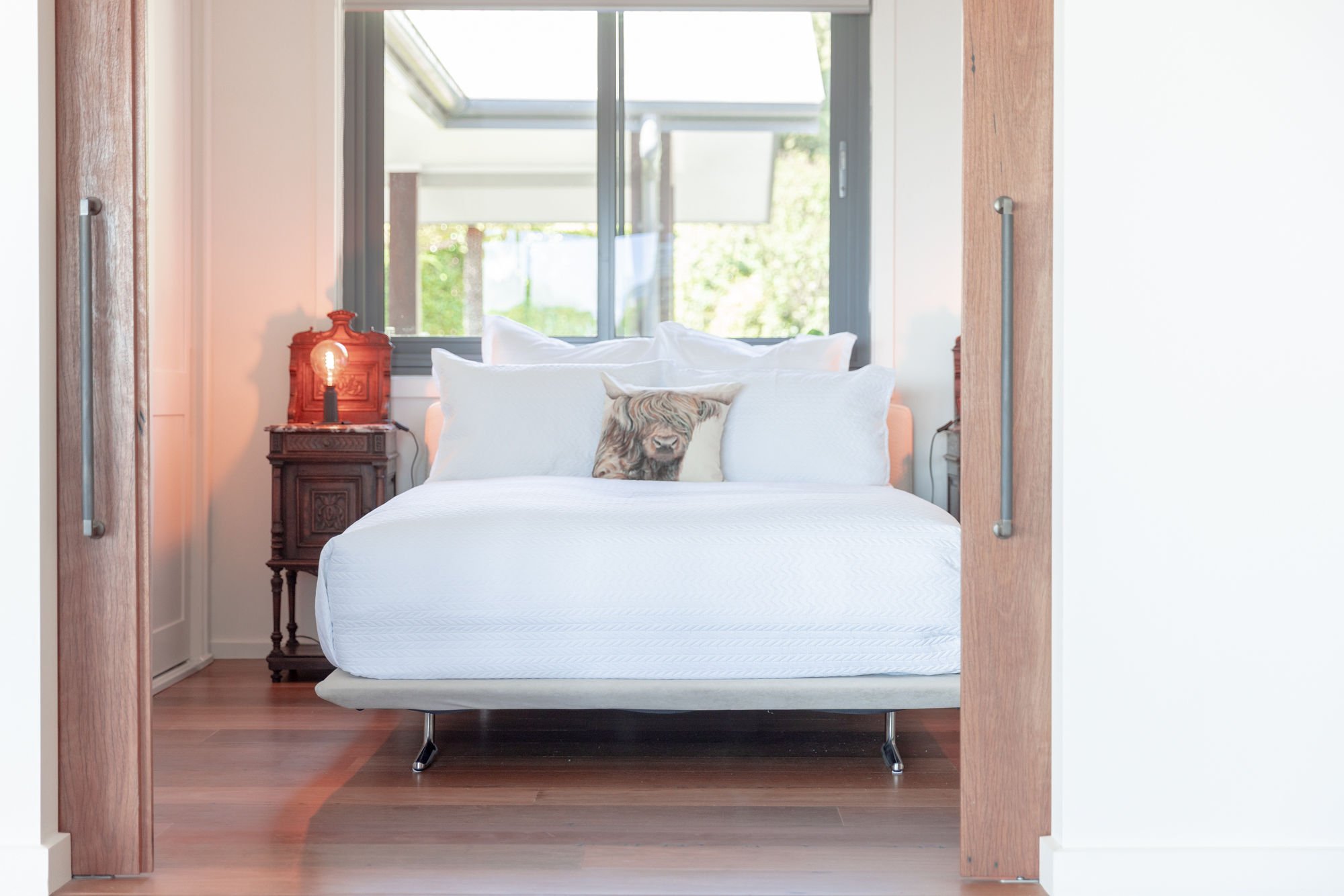 A modern bedroom with a bed, decorative pillows including one with a lion face, bedside table with a lamp, and large window showing trees outside.