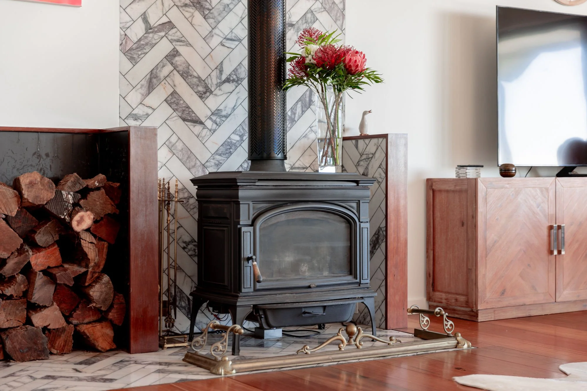 A fireplace in a living room with a black metal wood stove, decorated with a large glass vase of red and white flowers on top. To the left, there is a pile of firewood. The wall behind the fireplace features a geometric marble tile pattern, and to th