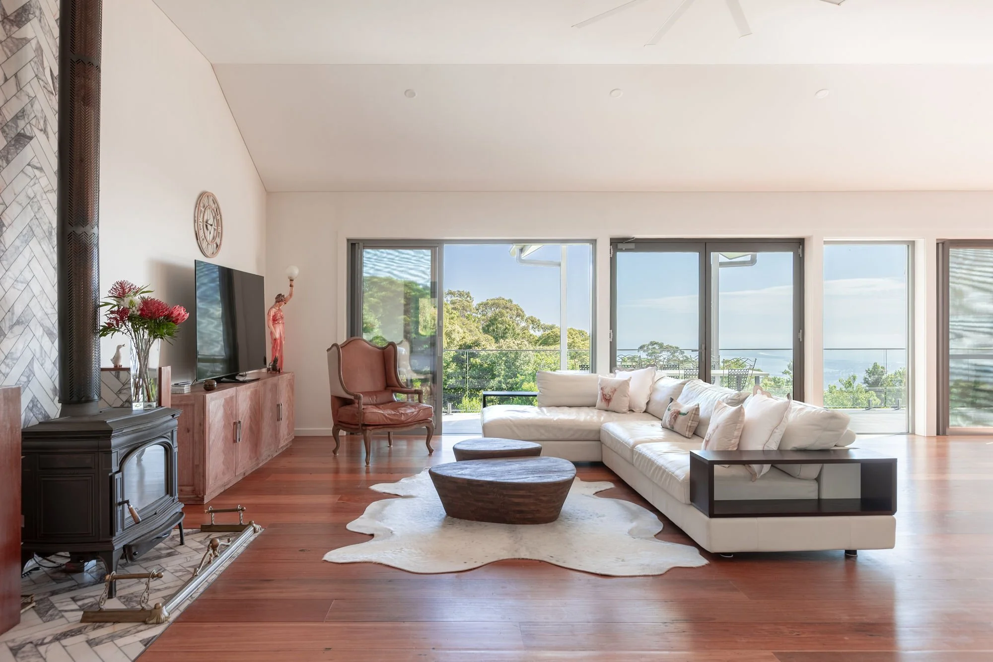 Living room with white sectional sofa, wooden coffee tables, pink armchair, fireplace, and large sliding glass doors leading to a balcony with trees and a view of the water.